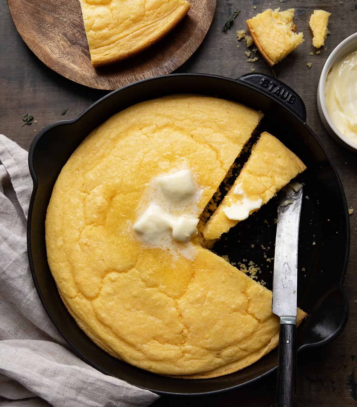 Skillet of Country Homestyle Cornbread on a wooden table from overhead with a few pieces cut out.