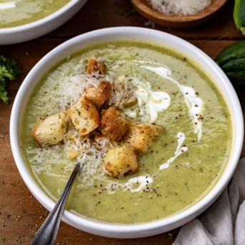 Bowl of Zucchini Soup with homemade croutons, parmesan cheese, and sour cream and a spoon in the bowl.