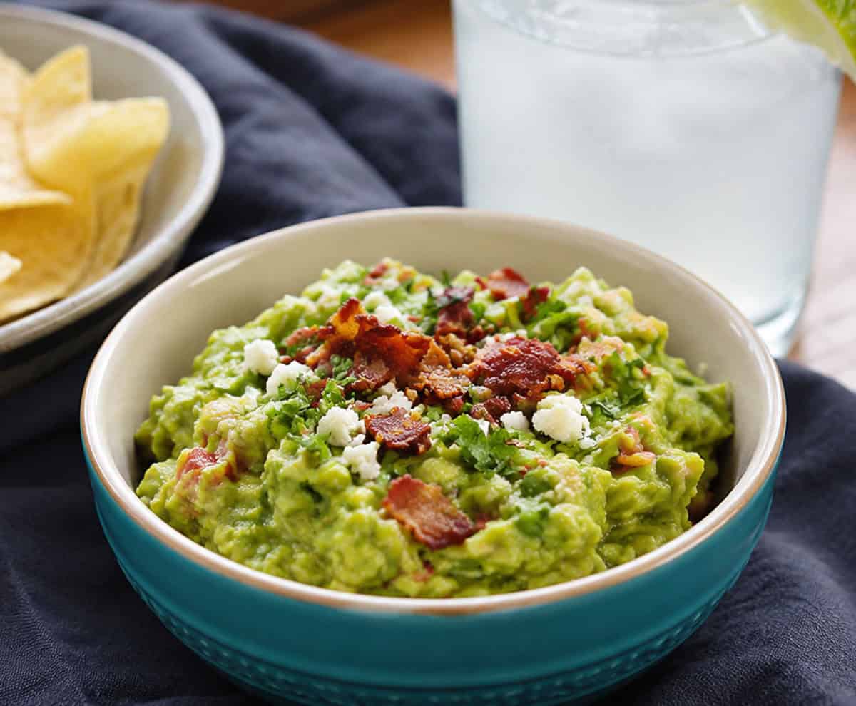 This isn’t your average guacamole! My Loaded Bacon Homemade Guacamole takes creamy avocado and fresh veggies and kicks it up with crispy bacon and all the vegetables! Bowl of loaded guacamole on a table with chips and a drink.