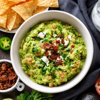 Bowl of Loaded Guacamole on a wooden table with crispy bacon, chips, avocado from overhead.