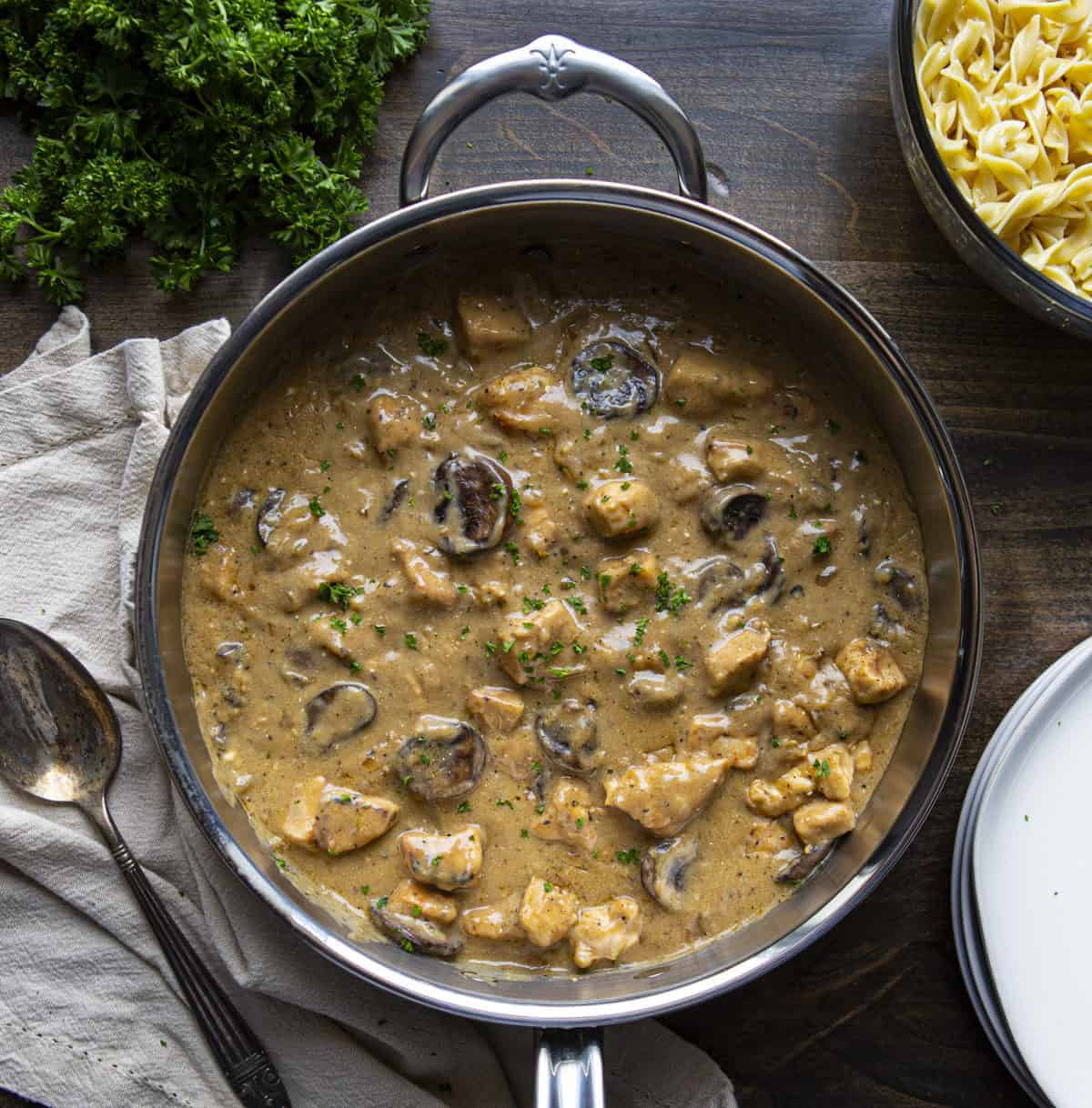 Overhead of skillet of Chicken Stroganoff Recipe with noodles, parsley, and plates by it