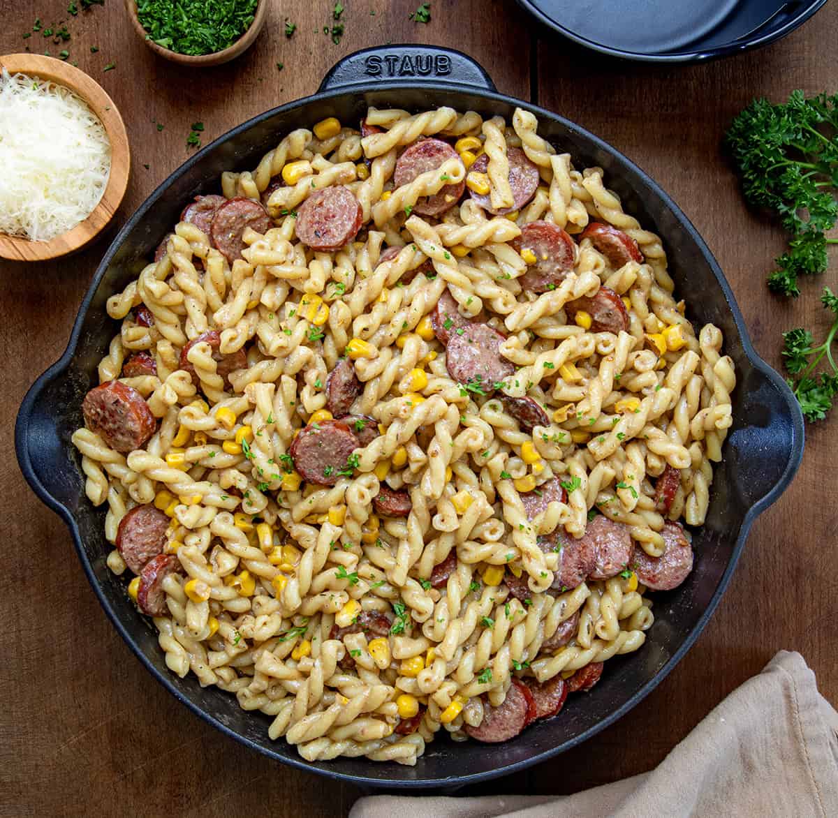 Smoked Sausage Pasta is a completely well-rounded and delicious meal filled with smoked sausage, corn, parmesan cheese, pasta, and just the right amount of Cajun seasoning. It is easy to put together and get on your dinner table in less than 30 minutes! Skillet of Smoked Sausage Pasta on a wooden table from overhead.