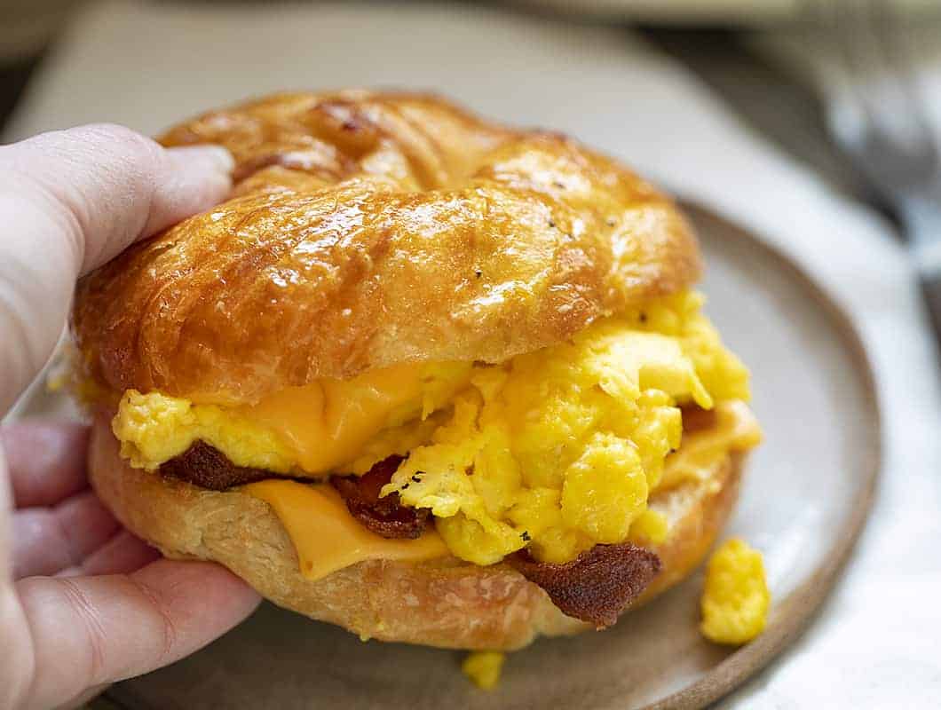 Hand Picking Up a Bacon Egg and Cheese Croissant Sandwich from Plate