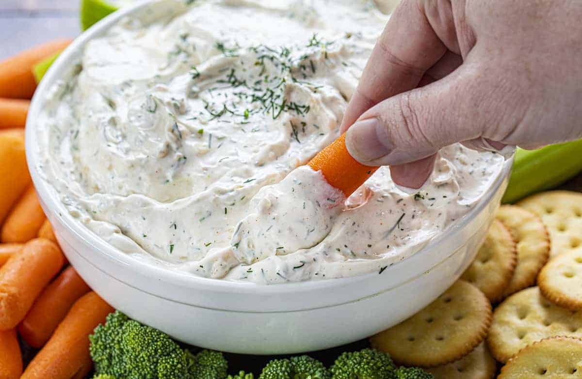 Hand Dipping Carrot into Dill Dip in White Bowl