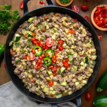 Skillet of Summer Goulash on a wooden table surrounded by fresh tomatoes and zucchini.