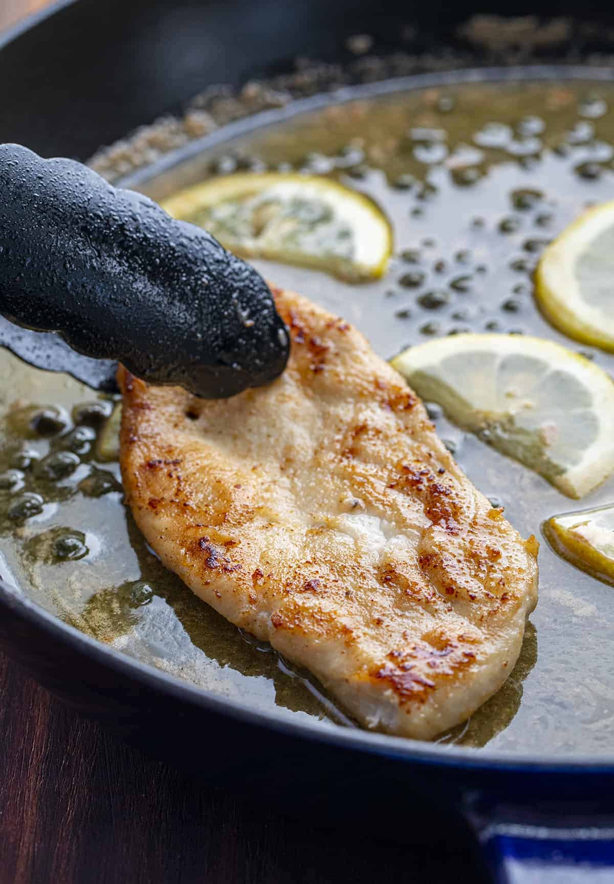 Tongs Holding a Piece of Chicken that is Being Laid in Sauce for Chicken Piccata 