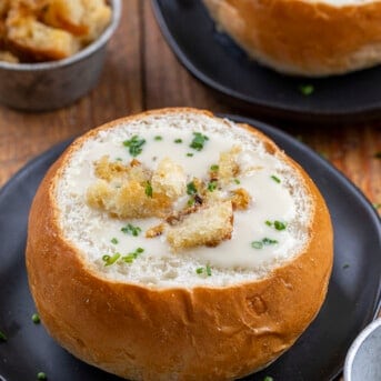 Bread Bowl Filled with 100 Clove Roasted Garlic Soup on a Black Plate with More Soup in the Background.