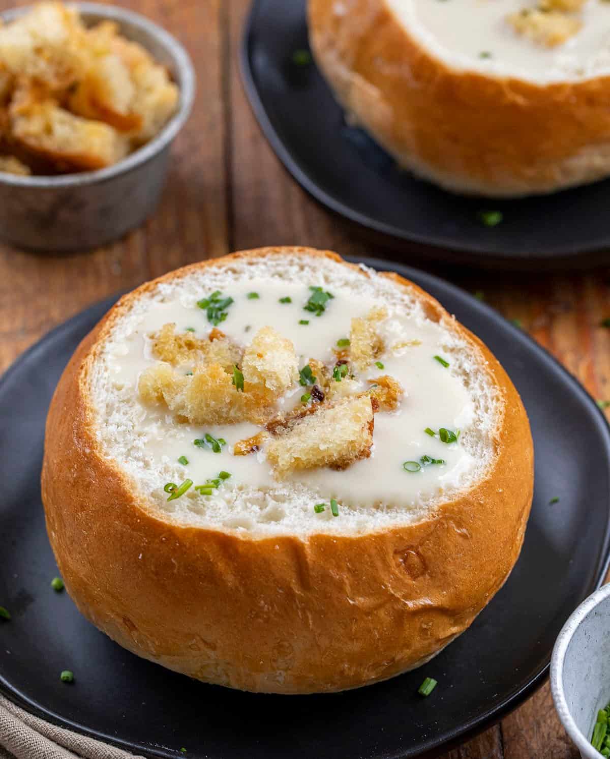 Bread Bowl Filled with 100 Clove Roasted Garlic Soup on a Black Plate with More Soup in the Background.