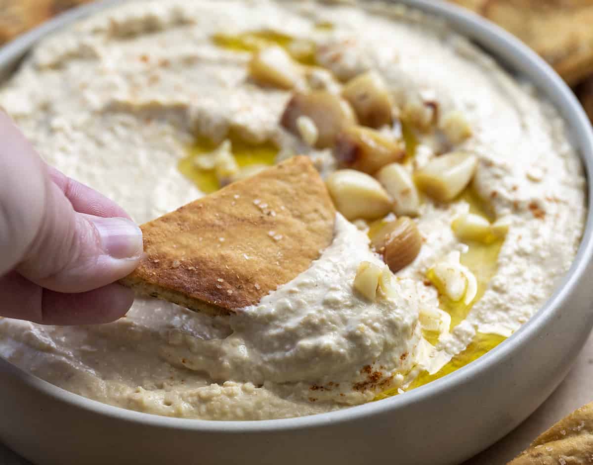 Dipping a Pita Chip into Homemade Roasted Garlic Hummus. Hummus, Hummus Recipes, How to Make Creamy Hummus, Ice Cubes in Hummus, Appetizer, What to Eat with Hummus, recipes, i am homesteader, iamhomesteader