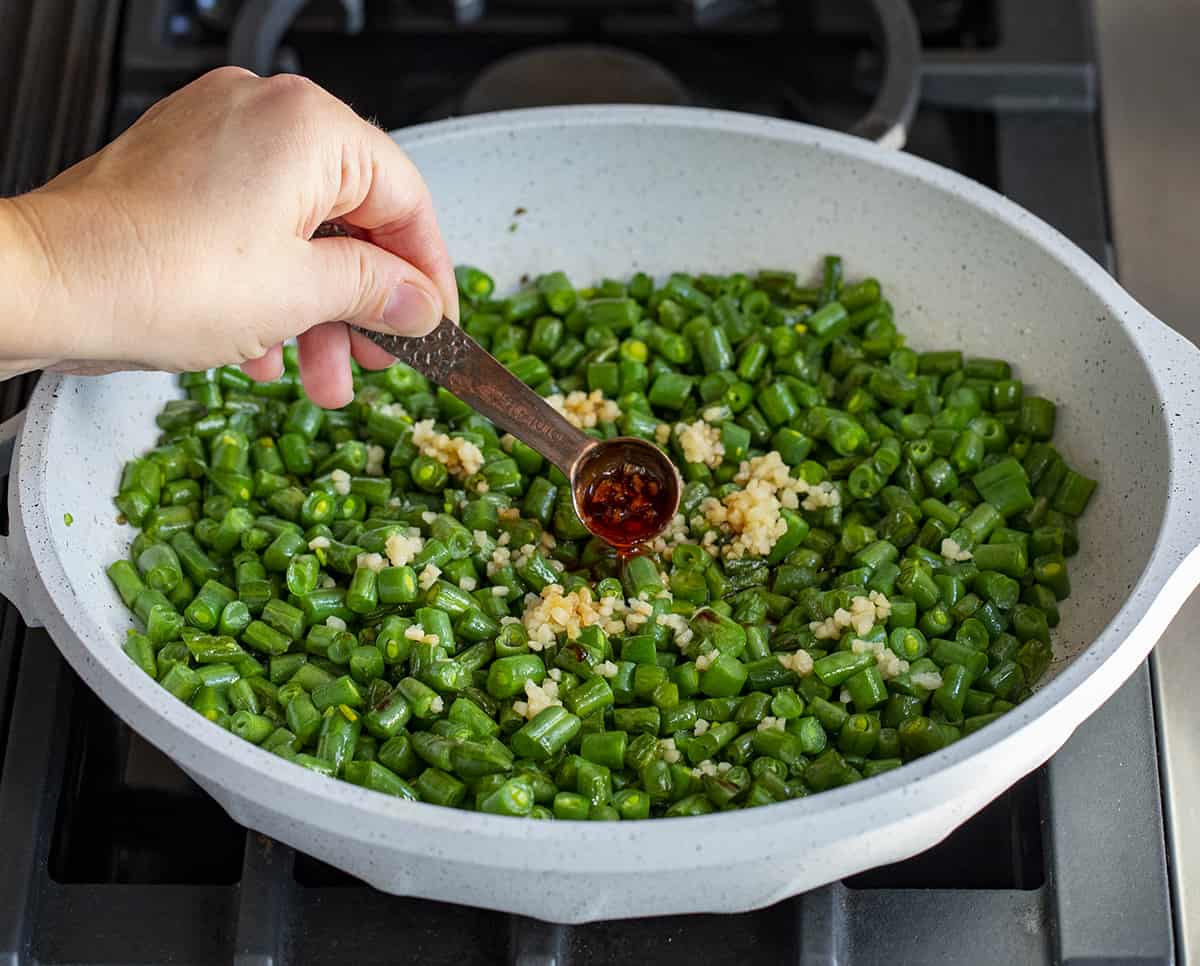 Adding Garlic Chili Sauce to Chopped Garlic Chili Green Beans in a Pan on a Stovetop.