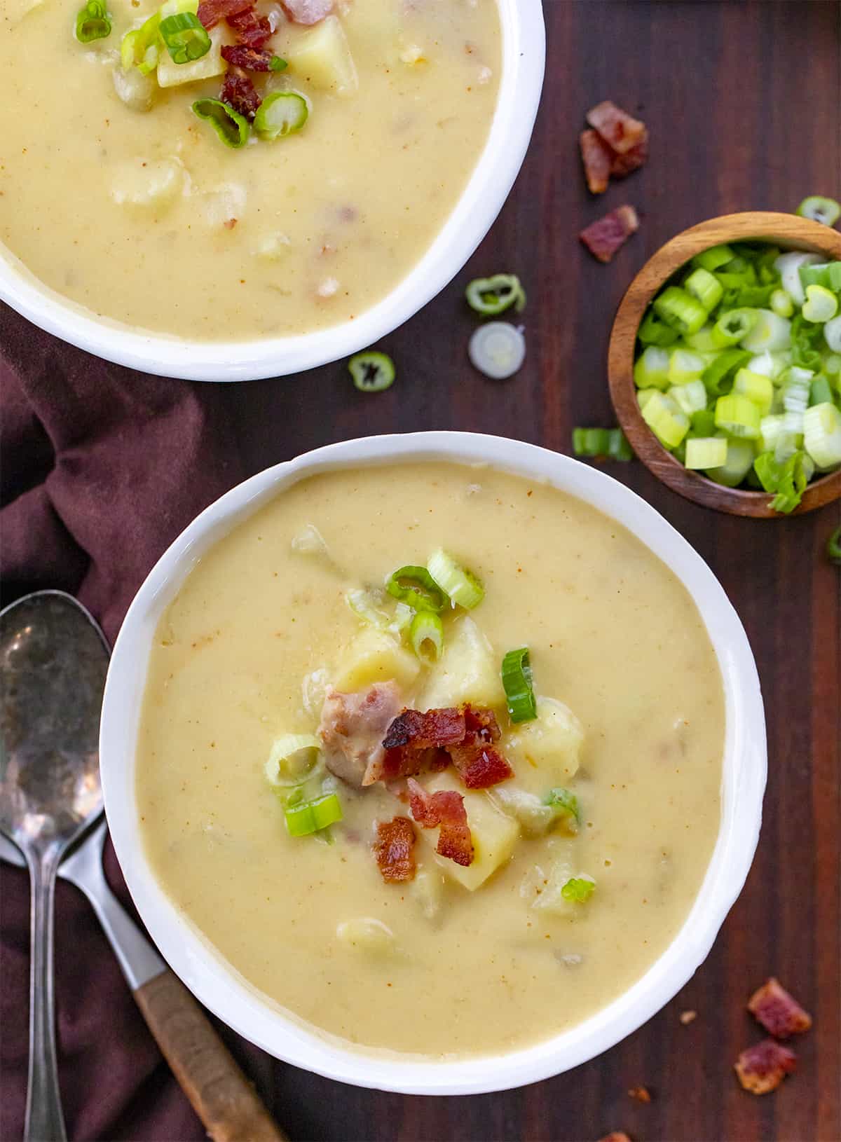 Two bowls of potato soup on a cutting board with green onion and bacon garnish and more green onion on the side. 