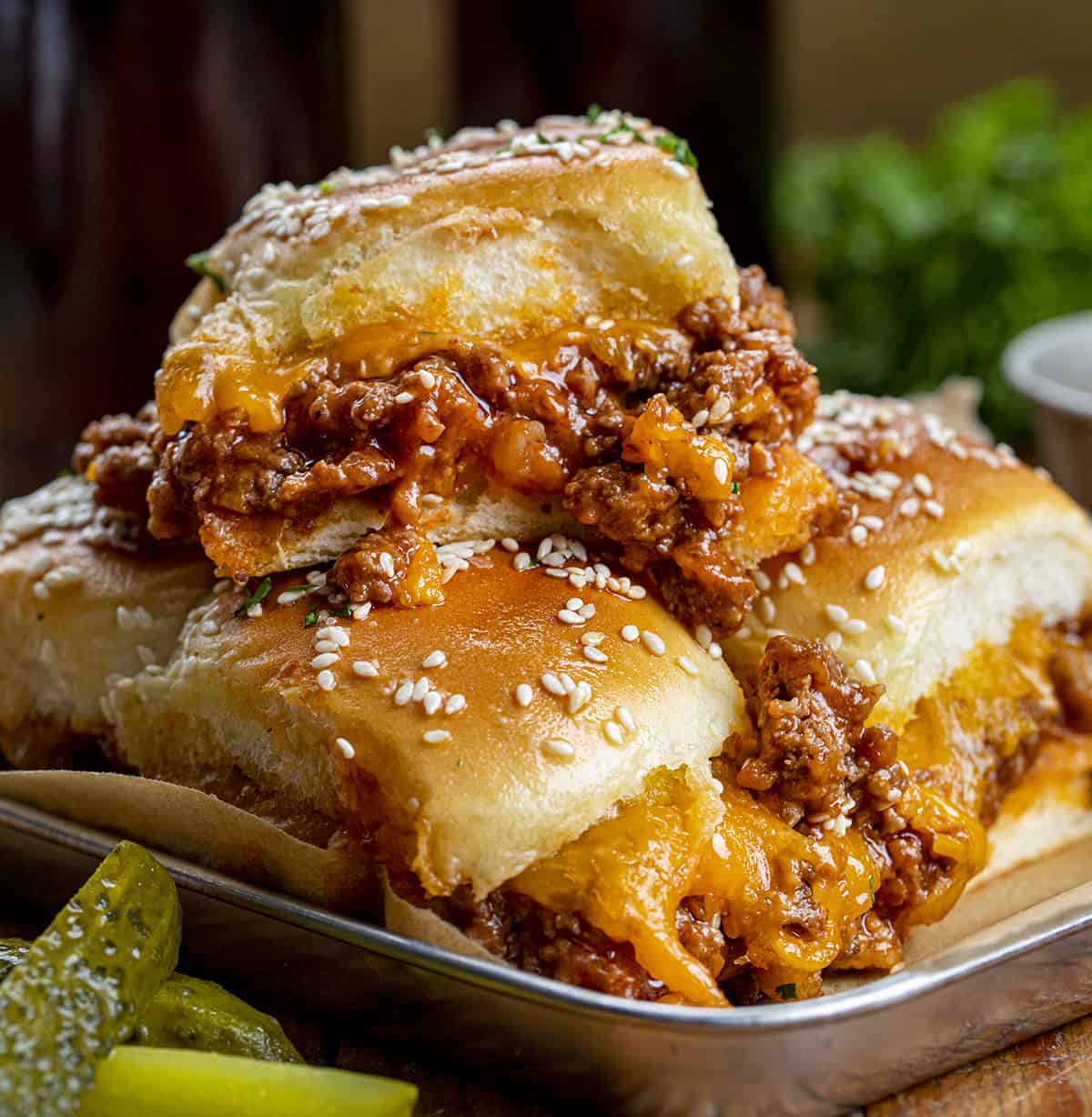 Sliders are one of the easiest ways to feed a crowd. They’re fun, customizable, and perfect for parties, game days, holidays, and family dinners! Sloppy Joe Sliders on a Tray with pickles and Beef in the Background.