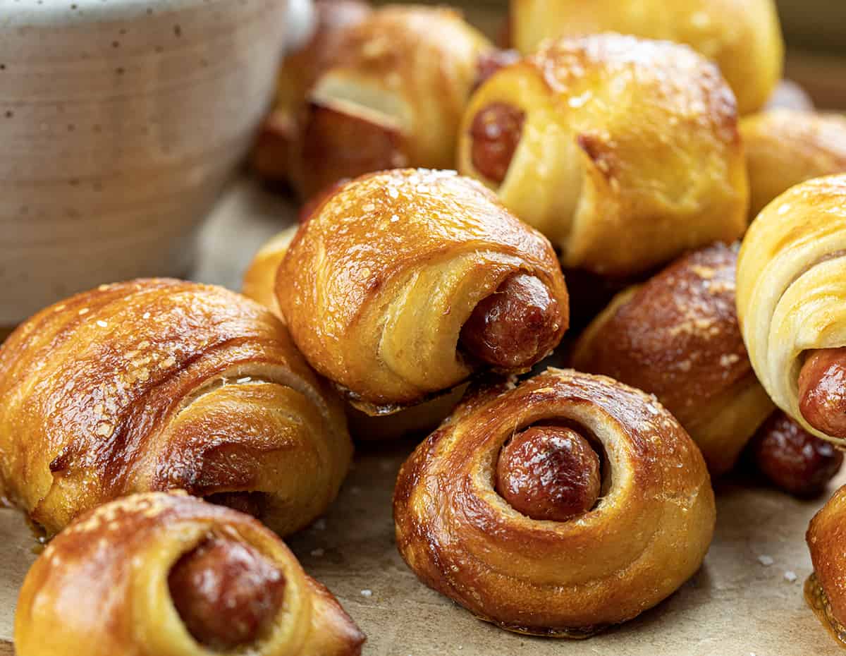Close up of Pretzel Pigs in a Blanket on a Cutting Board Next to a Bowl of Cheese Dip.