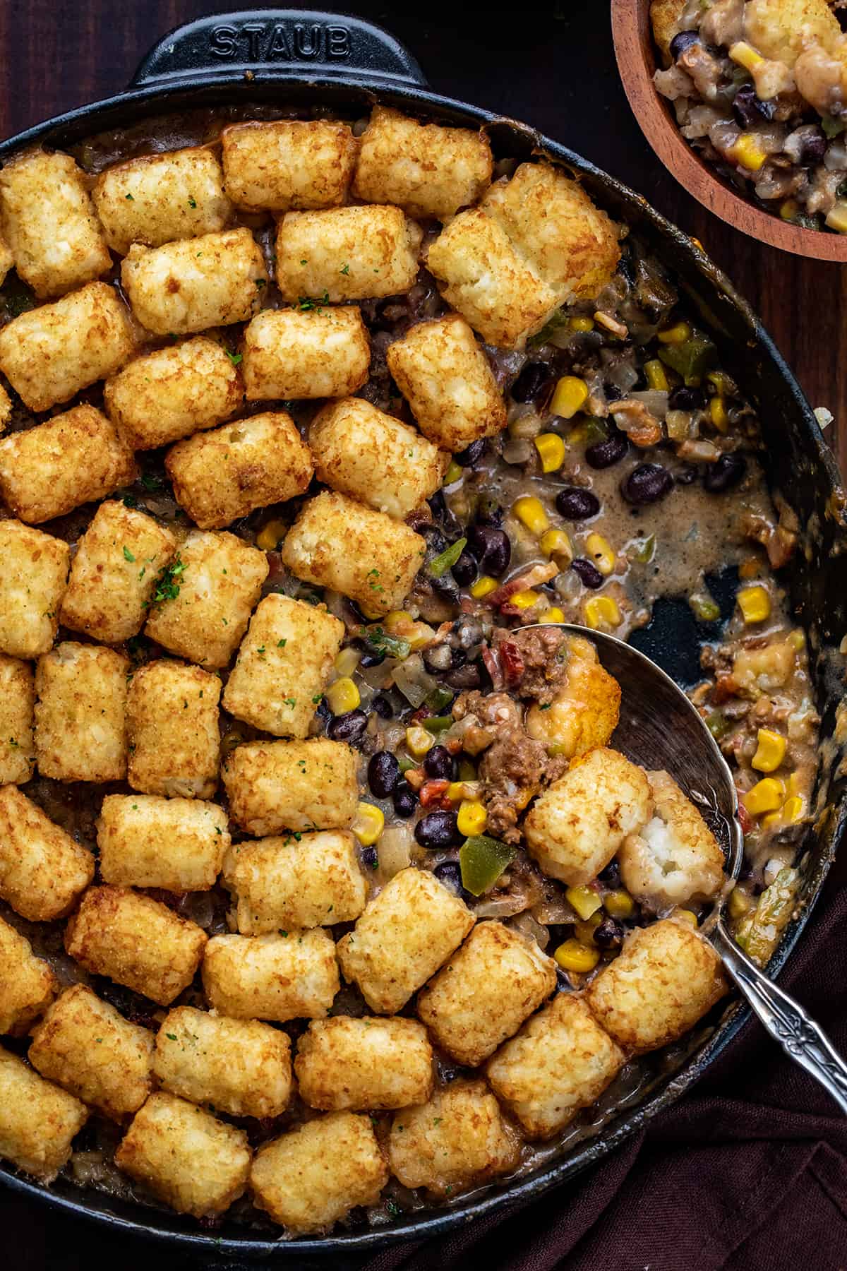 Skillet of Cowboy Tater Tot Casserole with Some Removed in a Bowl and a Spoon Resting in the pan.