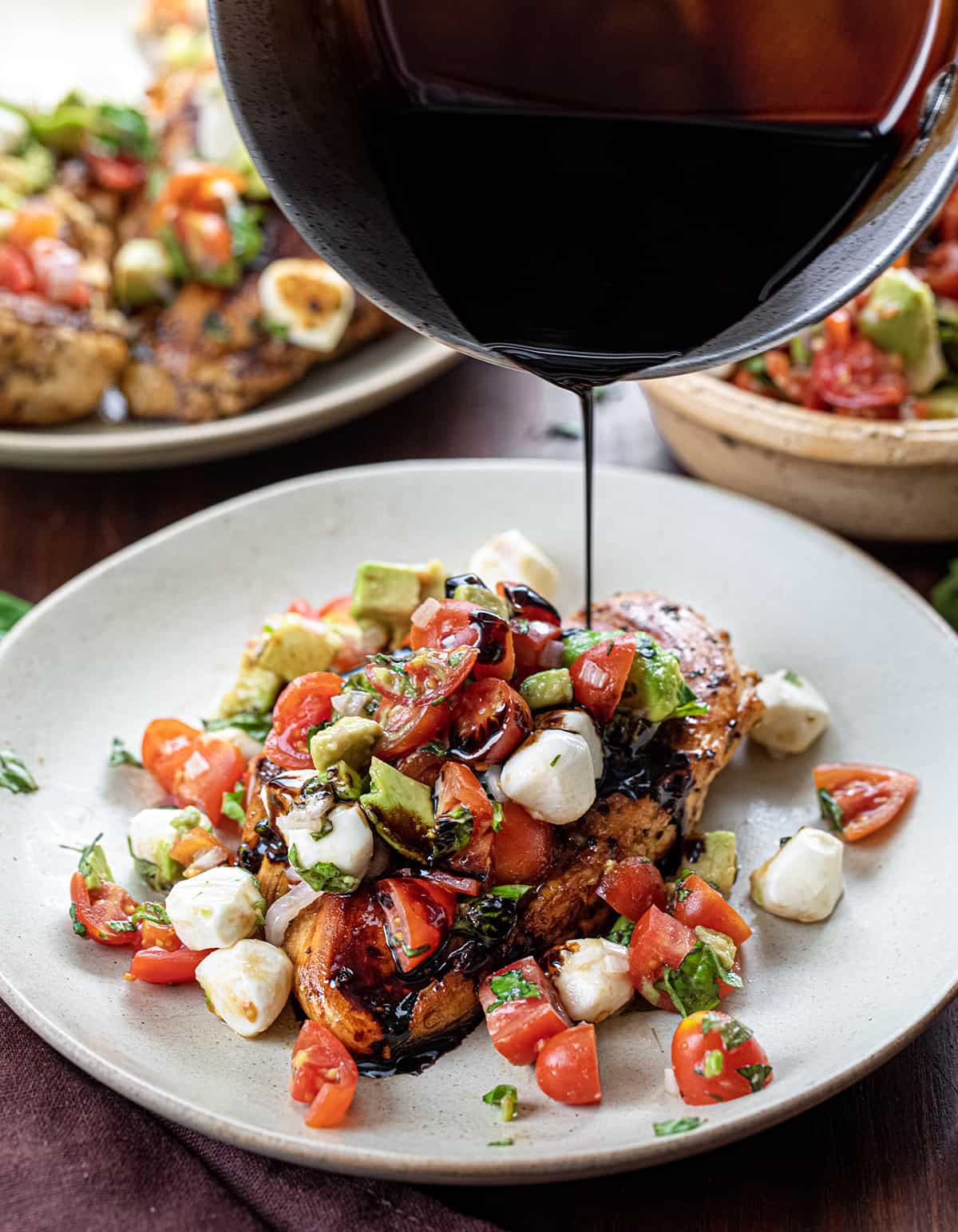 Pouring Balsamic Reduction Over Balsamic Chicken With Caprese Salsa on a Light Plate with More Caprese in the Back.