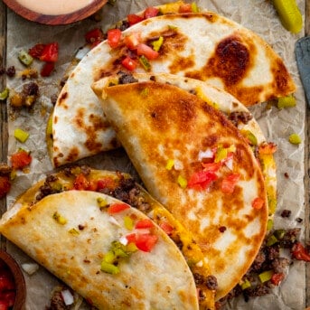 Three Cheeseburger Quesadillas on a Piece of Parchment with Tomatoes and Pickles Around Them.