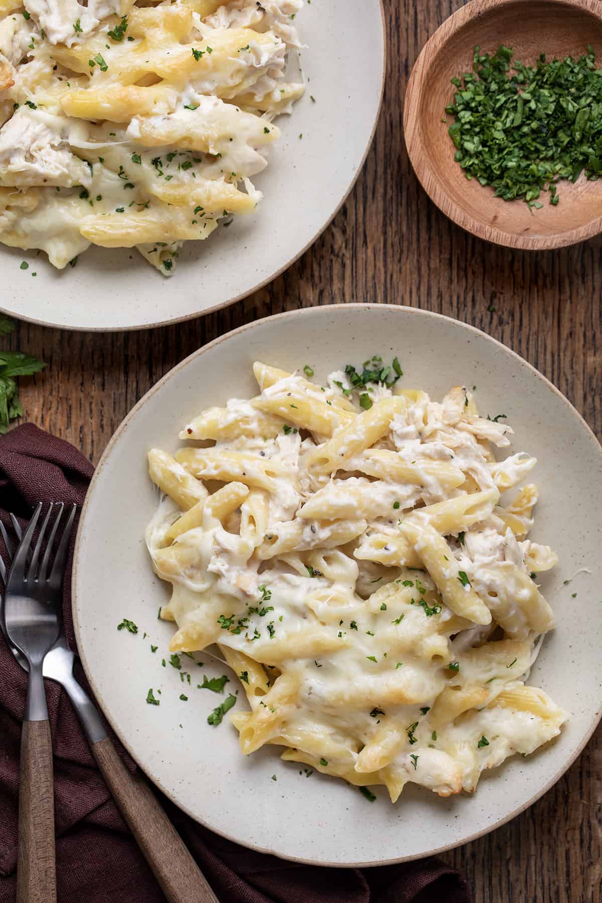 Two plates with Chicken Alfredo Bake on them on a Cutting Board with garnish and forks nearby.