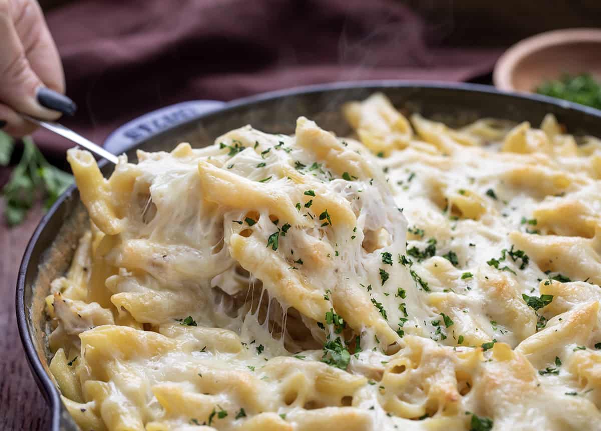 Spatula picking up a cheesy section of Chicken Alfredo Bake from a Skillet. 
