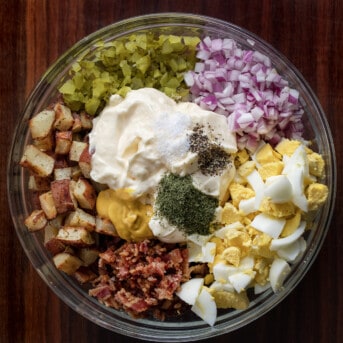 Raw Ingredients to Make Dill Pickle Roasted Potato Salad in a Glass Bowl on a Dark Cutting Board.