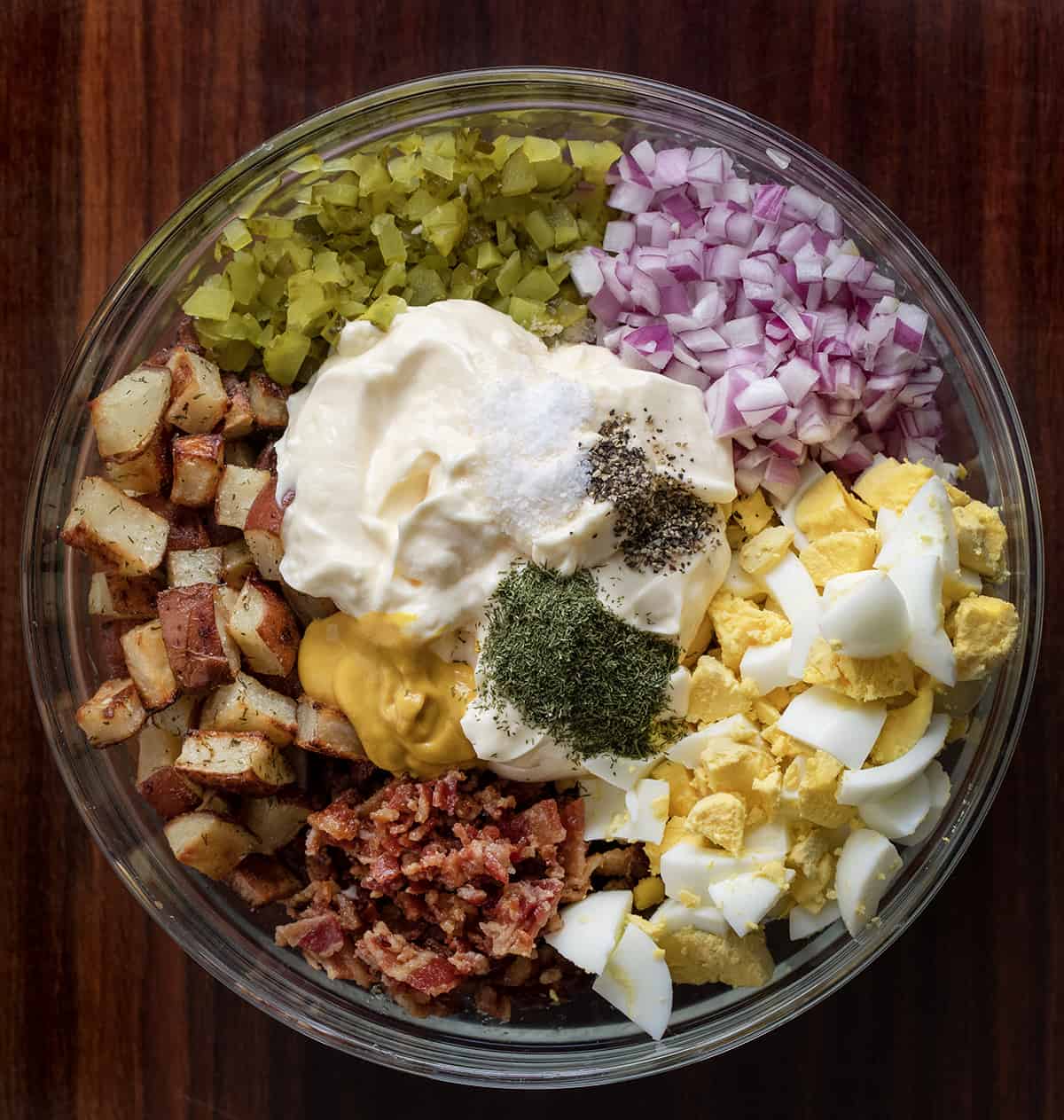 Raw Ingredients to Make Dill Pickle Roasted Potato Salad in a Glass Bowl on a Dark Cutting Board.