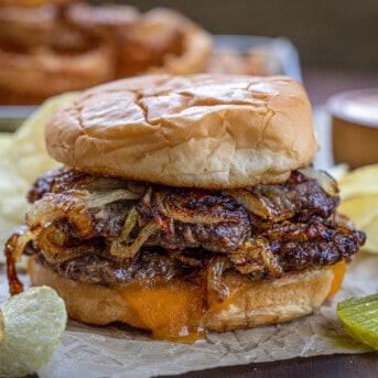 Oklahoma Fried Onion Burger on Parchment Paper with Pickles, Cheese, and Onion Rings in the Background.