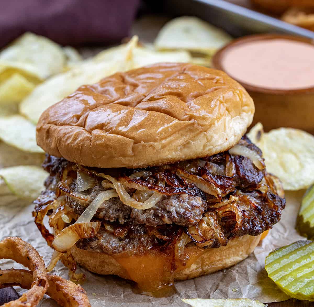 Oklahoma Fried Onion Burger on Parchment Paper with Pickles, Cheese, and Onion Rings in the Background.