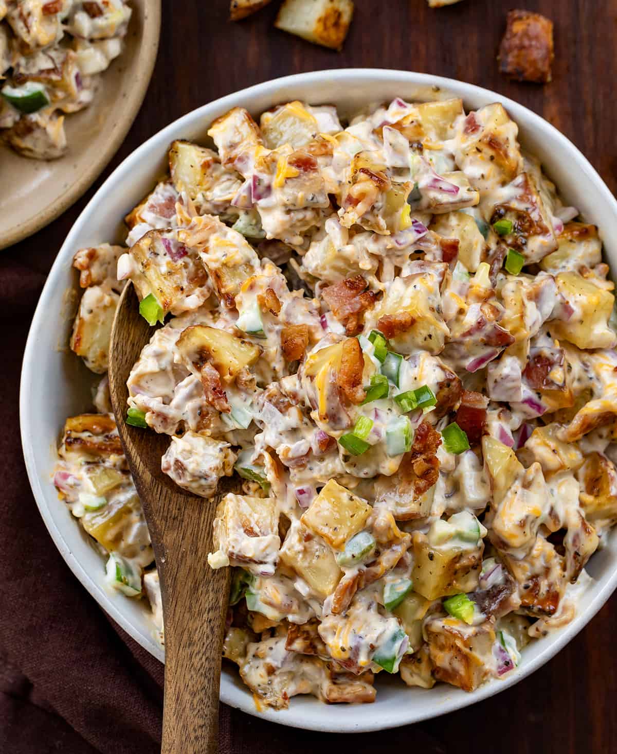 Bowl of Jalapeño Popper Roasted Potato Salad on a Dark Cutting Board with Wooden Spoon in Bowl.