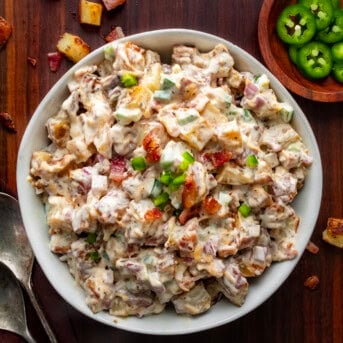 Bowl of Jalapeño Popper Roasted Potato Salad on a Dark Cutting Board with Spoons and Jalapenos.