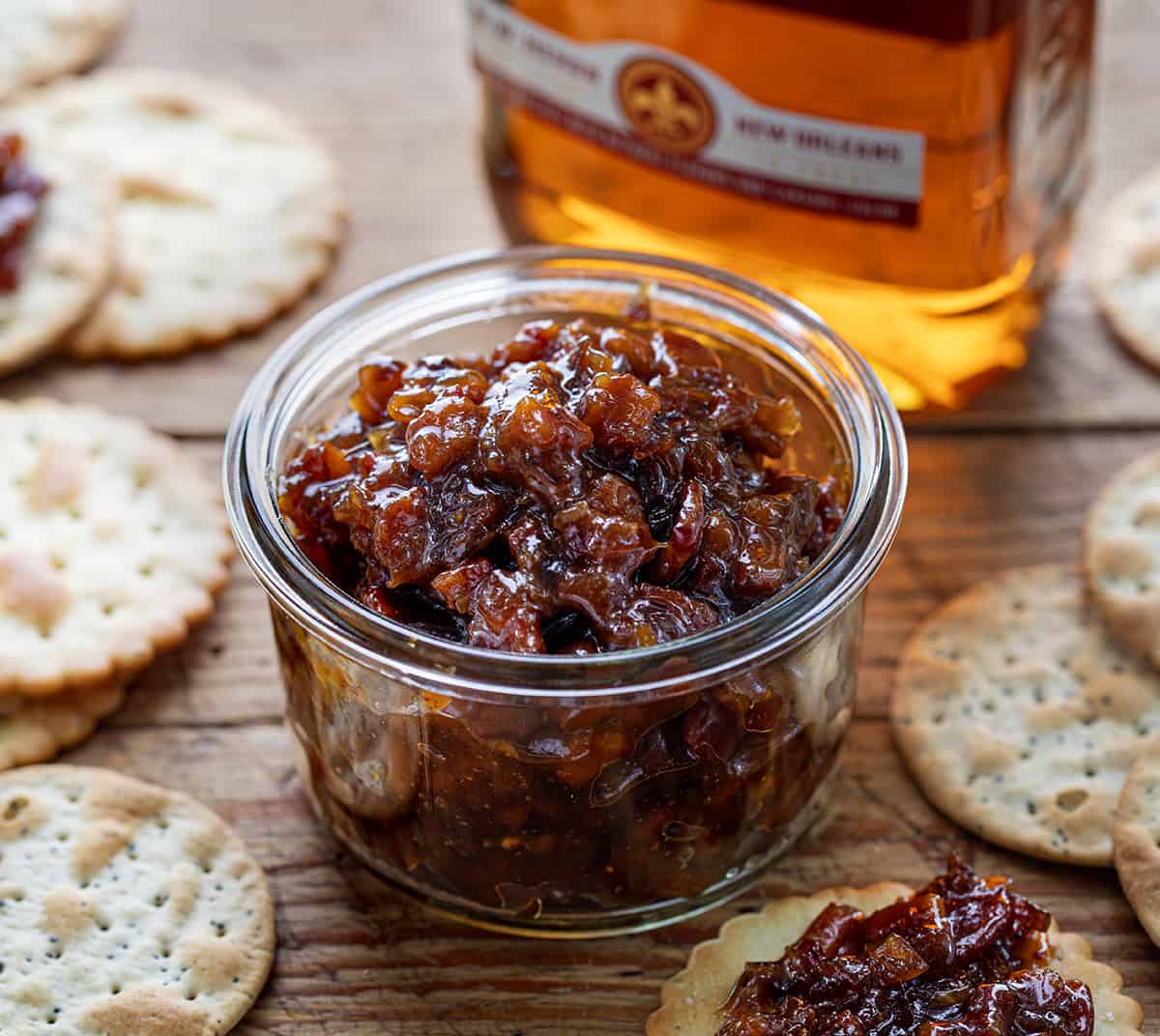Jar of Whiskey Bacon Jam on a Cutting Board with Crackers and Whiskey in the Background.