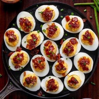 Platter of Whiskey Bacon Jam Deviled Eggs with Chive Garnish on a Dark Cutting Board.