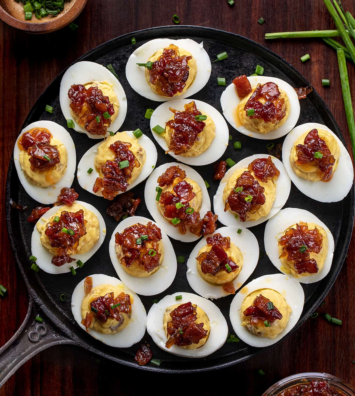 Platter of Whiskey Bacon Jam Deviled Eggs with Chive Garnish on a Dark Cutting Board. 