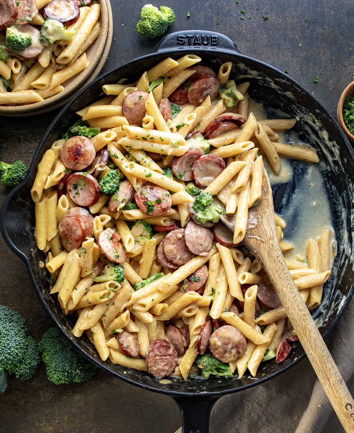 Creamy Kielbasa Broccoli Skillet on a Dark Table with Some Removed and Plated and a Wooden Spoon in the Pan.