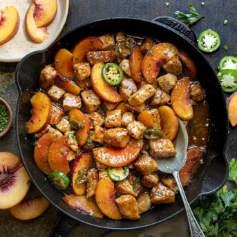 Skillet of Jalapeno Peach Pork Bites on a Dark Table with Fresh Peaches from Overhead.