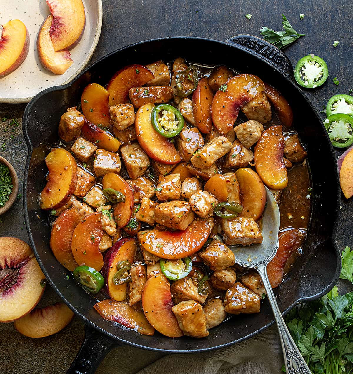 Skillet of Jalapeno Peach Pork Bites on a Dark Table with Fresh Peaches from Overhead. 