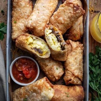 Steak Egg and Cheese Egg Rolls in a Pan with One Cut Open Showing the Steak and Eggs Inside on a Wooden Table.