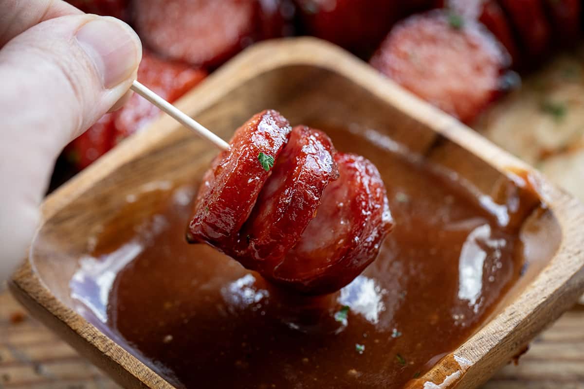 Hand Holding Maple BBQ Hasselback Kielbasa Bite on a Toothpick and Dipping in BBQ.