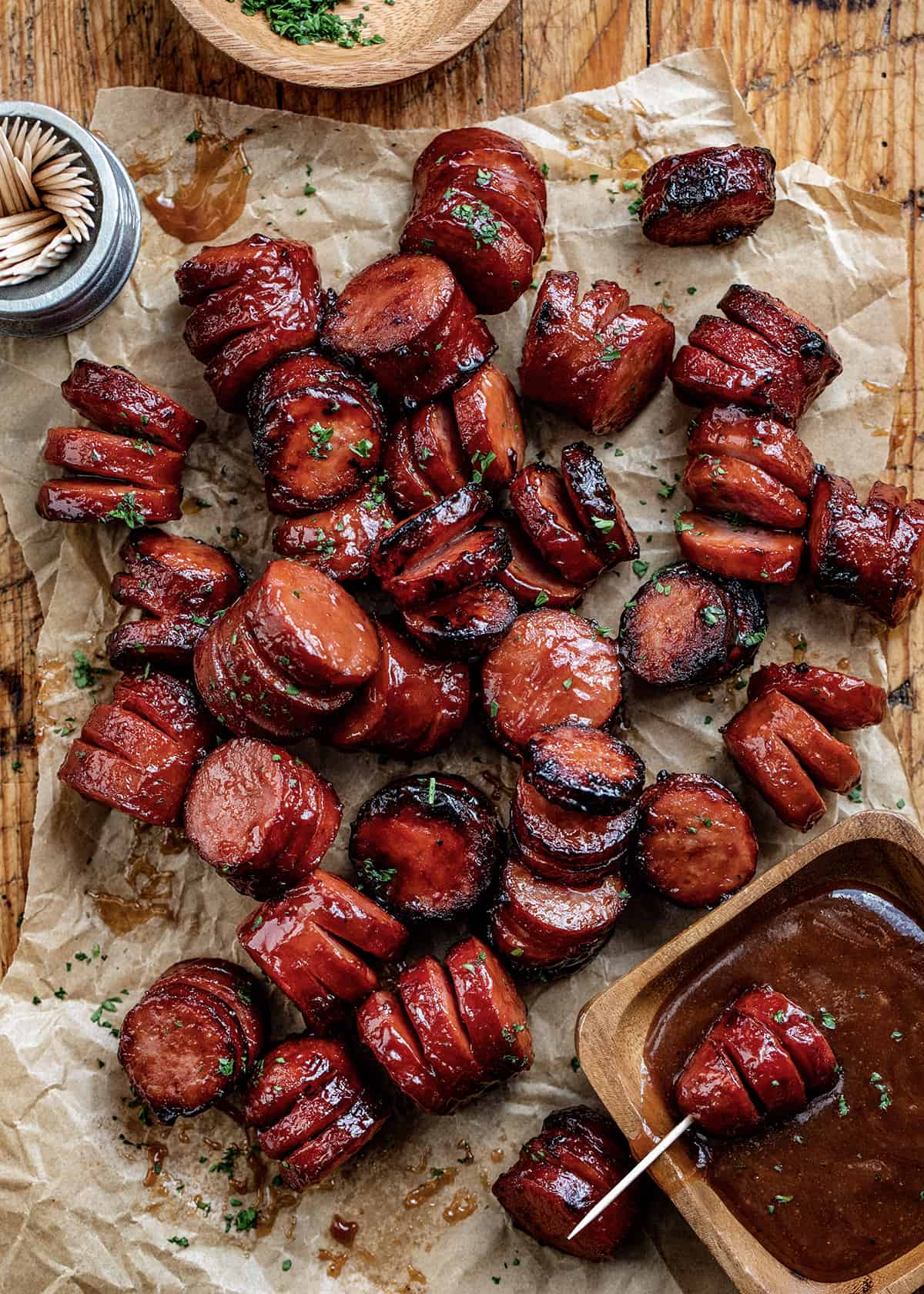 Looking Down on Maple BBQ Hasselback Kielbasa Bites on Parchment Paper Next to BBQ Sauce and Toothpicks. 