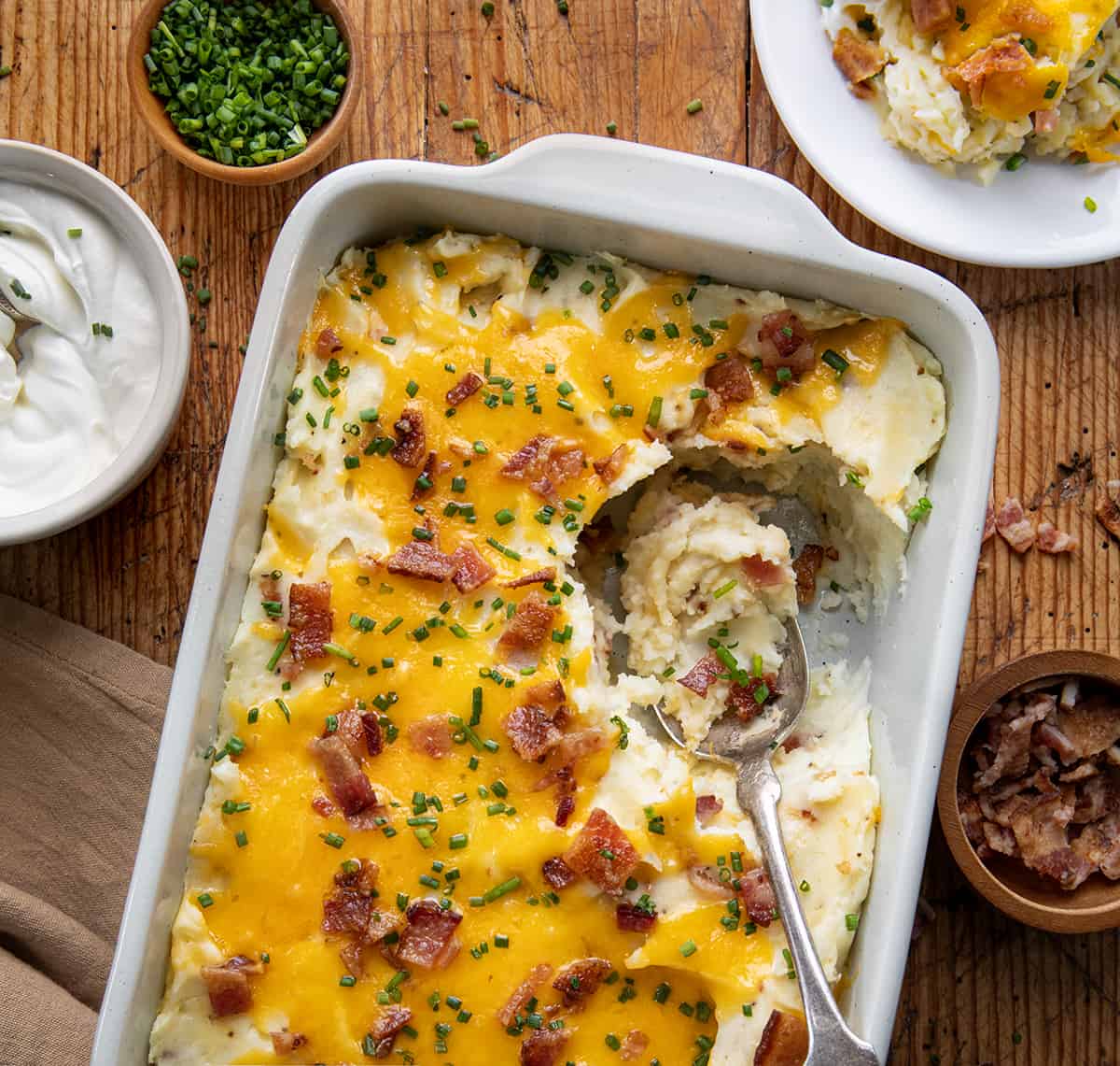 Pan of Loaded Mashed Potatoes with some removed and plated and then a spoon in the pan.