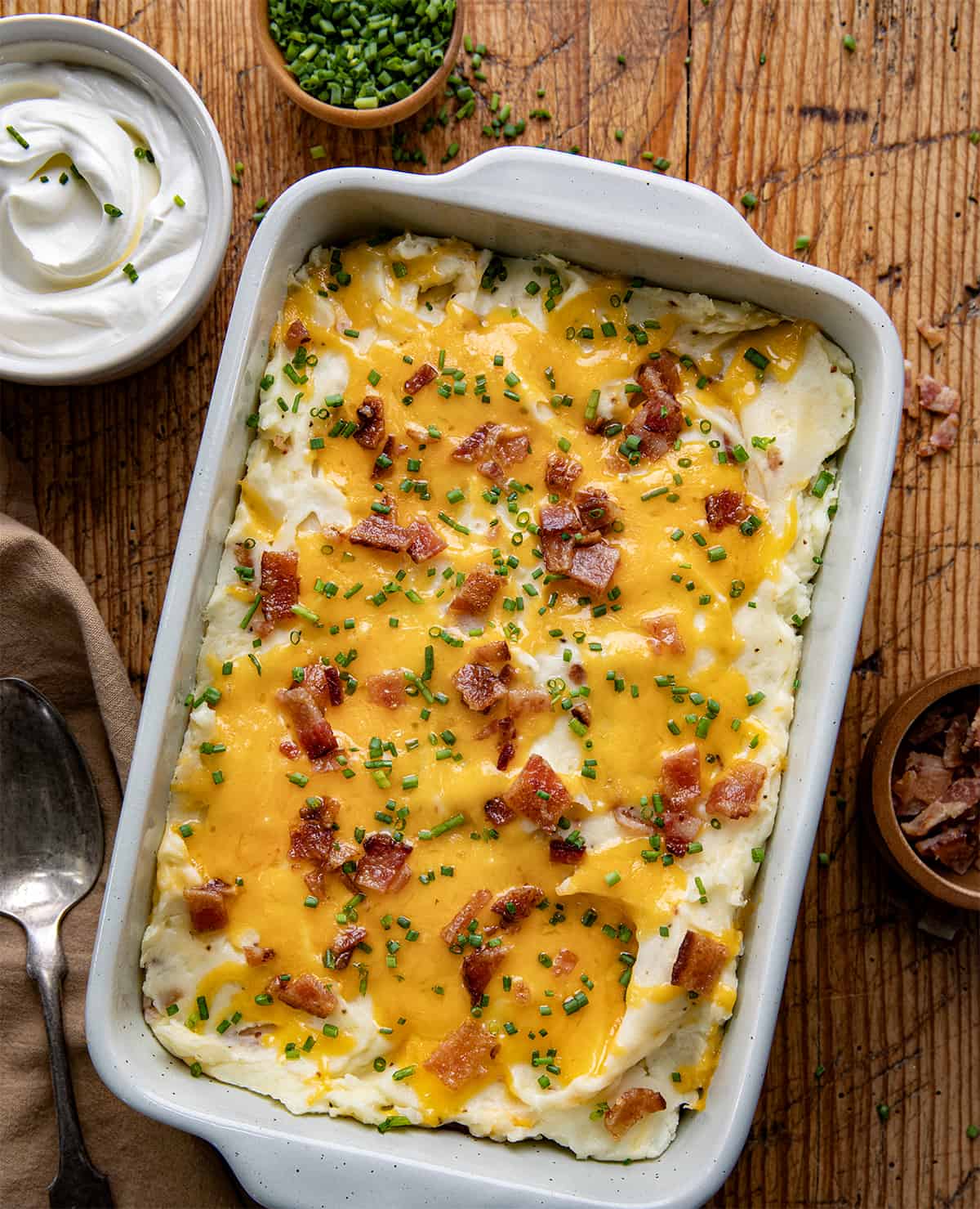 Pan of Loaded Mashed Potatoes on a wooden table next to sour cream and bacon and a spoon from overhead.