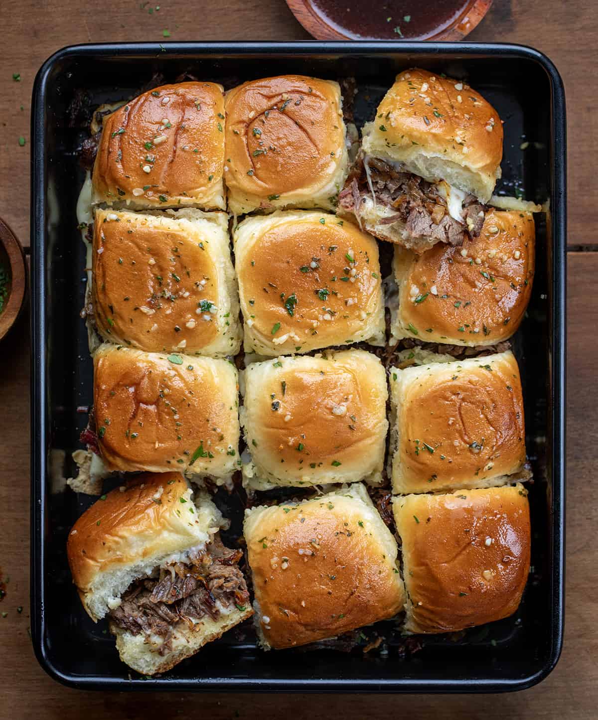 Pan of French Dip Sliders with some sliders on their side on a wooden table from overhead. 