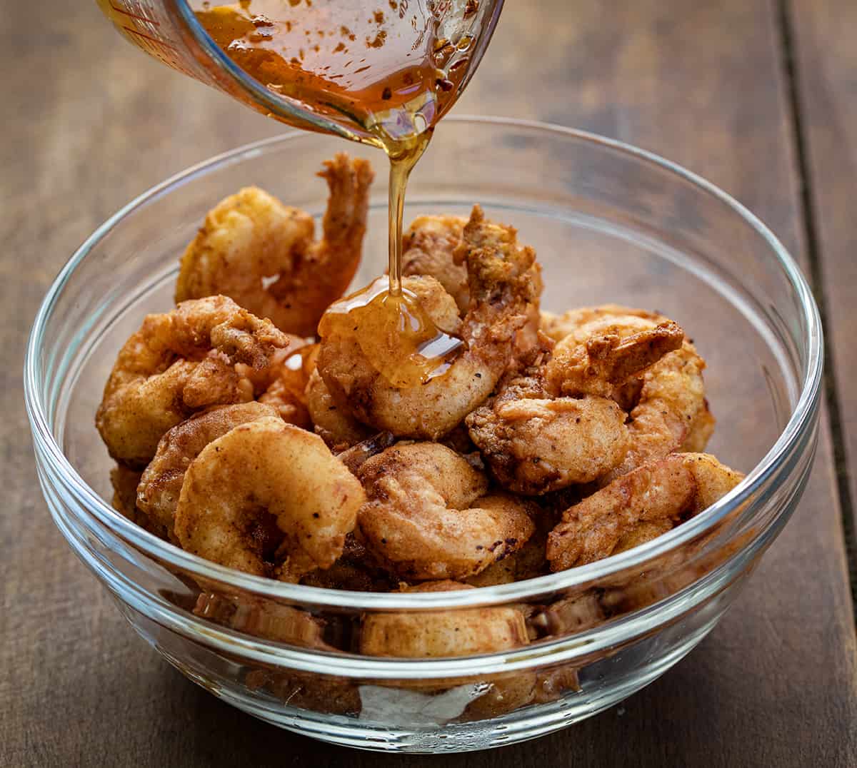 Pouring hot honey over fried shrimp in a glass bowl.