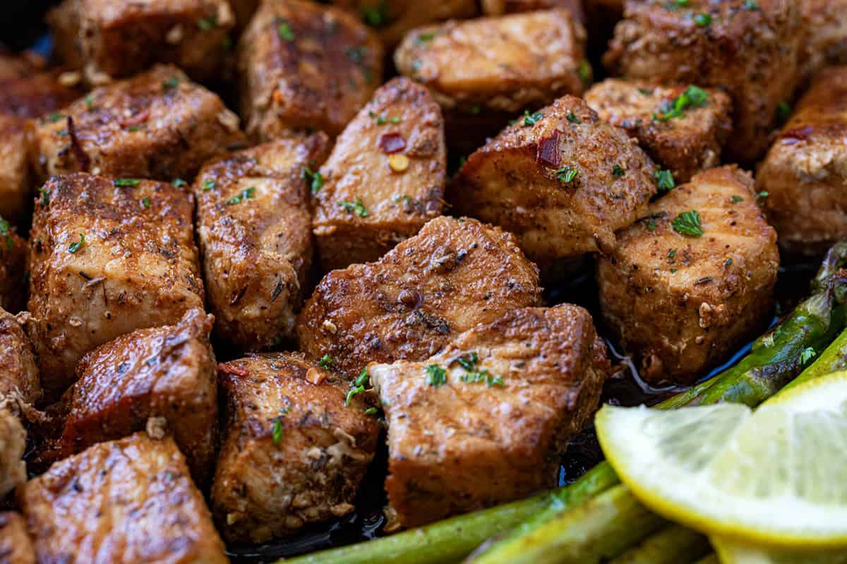 CLose up of blackened pork bites in a pan nestled next to asparagus and a lemon slice.