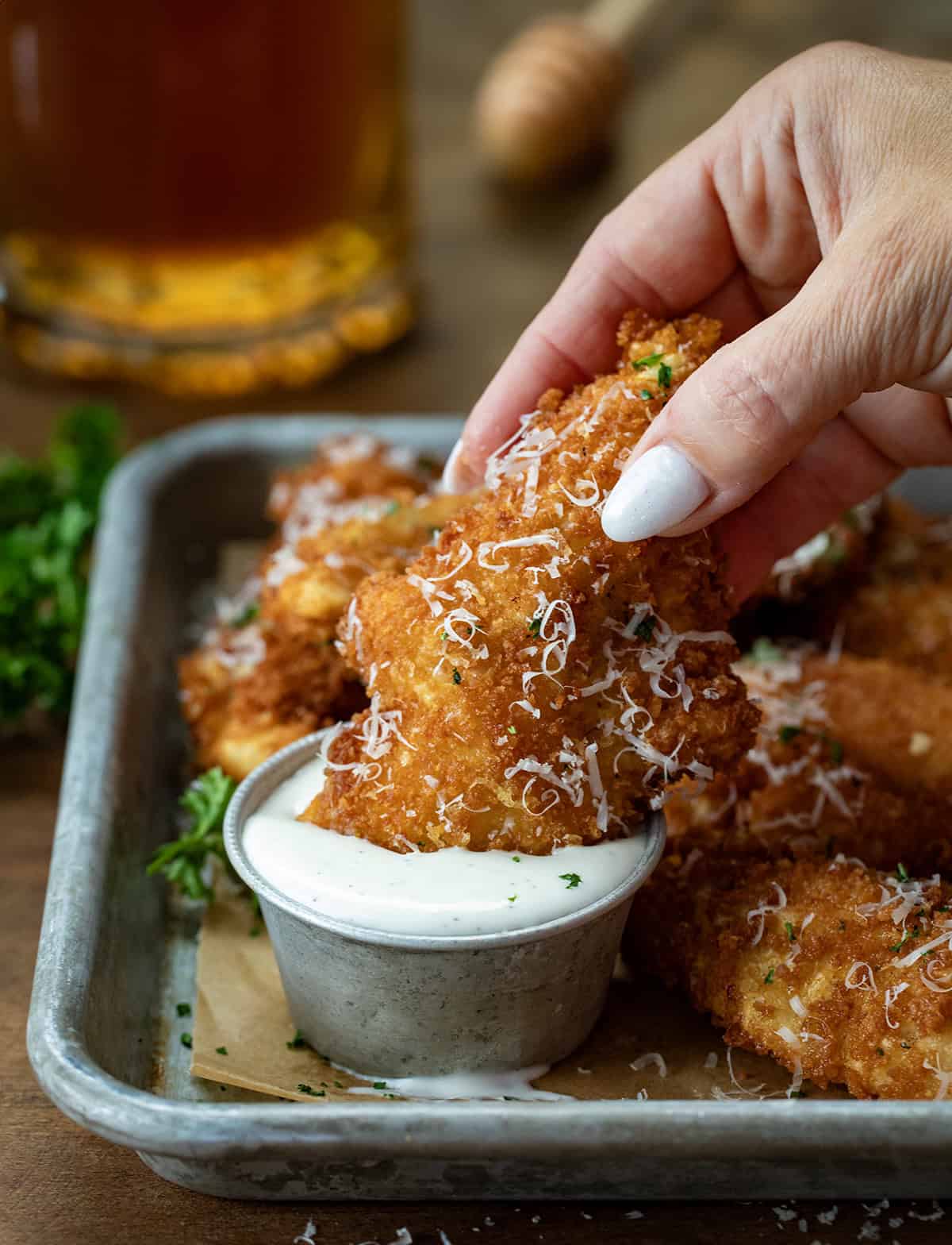 Hand dipping a Garlic Parmesan Chicken Tender into a bowl of ranch dressing on a wooden table.