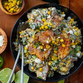 Mexican Street Corn Smashed Potato Salad in a black bowl on a wooden table with cojita cheese limes and corn around it.