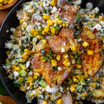 Mexican Street Corn Smashed Potato Salad in a black bowl on a wooden table with cojita cheese limes and corn around it.