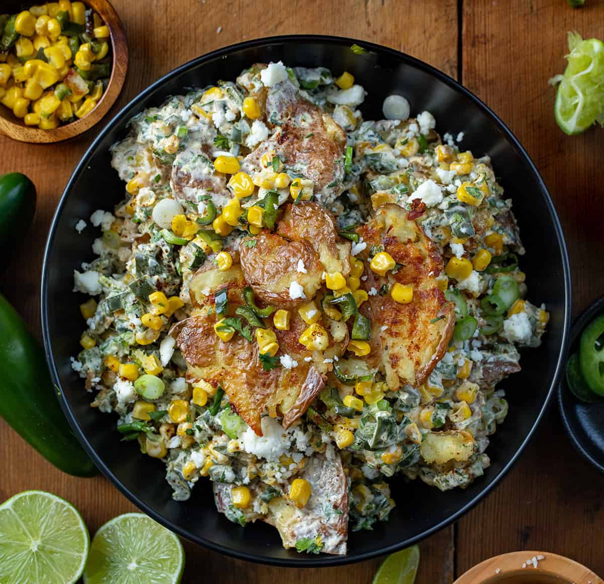 Mexican Street Corn Smashed Potato Salad in a black bowl on a wooden table with cojita cheese limes and corn around it. 