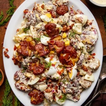 Platter of Smashed Roasted Potato Salad on a wooden table showing the smashed potatoes, hard boiled eggs, crispy bacon bits, finely diced onion, and perfect dressing that brings it all together.