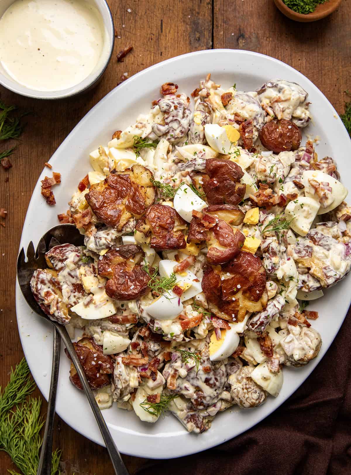 Platter of Smashed Potato Salad on a wooden table showing the smashed potatoes, hard boiled eggs, crispy bacon bits, finely diced onion, and perfect dressing that brings it all together.