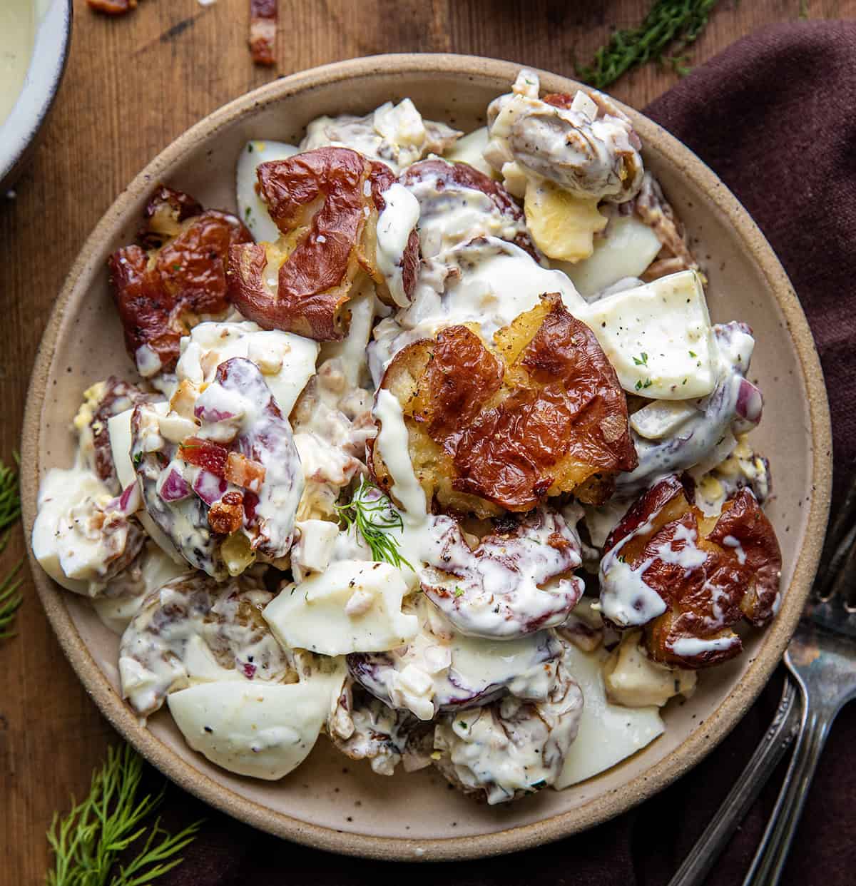 Plate of Smashed Roasted Potato Salad on a wooden table showing the smashed potatoes, hard boiled eggs, crispy bacon bits, finely diced onion, and perfect dressing that brings it all together.