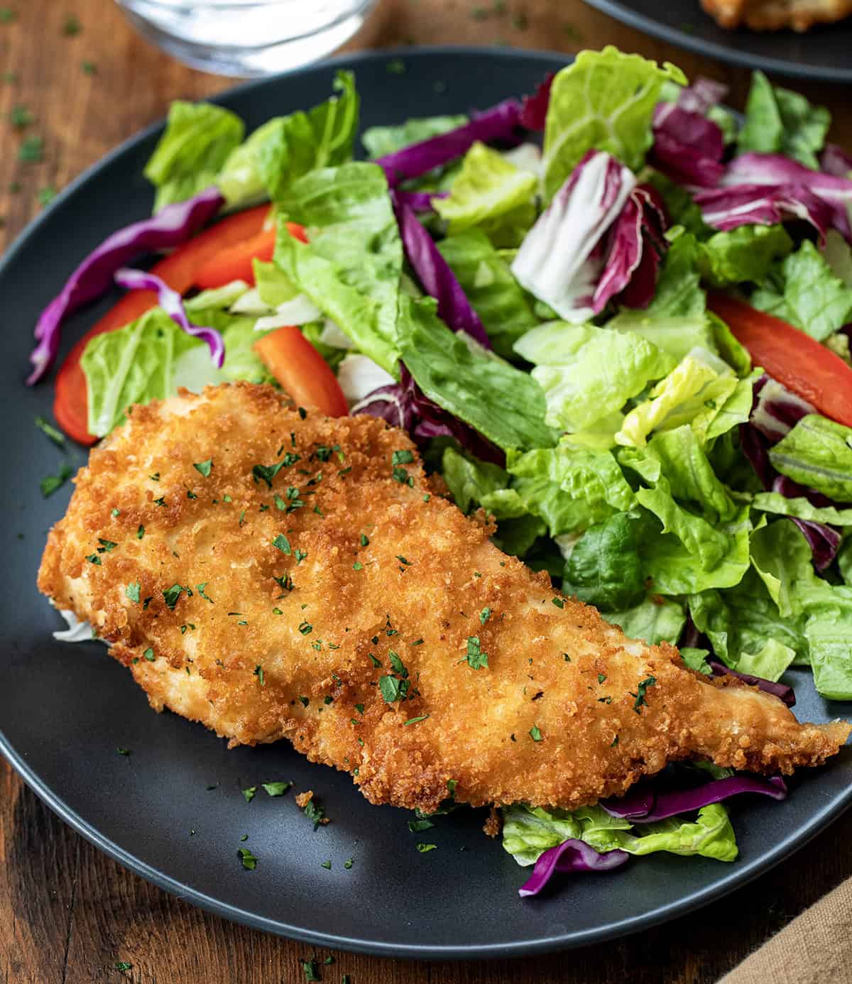 One chicken cutlet on a black plate with salad on a wooden table.